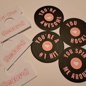 May include: A collection of black vinyl record-shaped cards with pink accents and white heart cutouts. Each card has a different phrase in pink, such as "You're Awesome" and "You Rock!" Also included are white card sleeves with the text "For the Record" in pink.