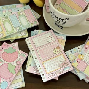 Könnte beinhalten: Eine Sammlung farbenfroher, dekorativer Notizblöcke mit verschiedenen Designs, darunter Erdbeeren, Katzen und pastellfarbene Rechtecke. Einige Notizblöcke befinden sich in einer weißen Tasse mit der Aufschrift "Paris".