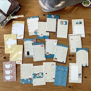 以下が含まれることがあります： 食べ物、飲み物、その他の可愛いイラストなど、様々なデザインが施された、18枚の印刷可能なプランナー用ステッカーのセット。ステッカーは白地に茶色の背景で印刷されており、上部に穴が開いています。