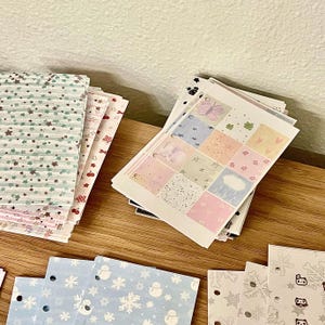 Könnte beinhalten: Eine Sammlung dekorativer Papierbögen mit verschiedenen Mustern, darunter Sterne, Schneeflocken und Schmetterlinge. Die Bögen weisen Pastellfarben auf und sind auf einer Holzoberfläche angeordnet, ideal für Bastelarbeiten und Scrapbooking.