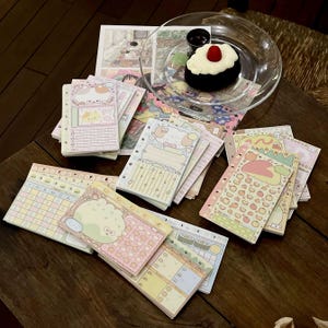 May include: A collection of pastel-colored, illustrated notepads with various designs, including cute animal characters and decorative elements. The notepads are arranged on a wooden surface, alongside a small dessert and a comic strip.