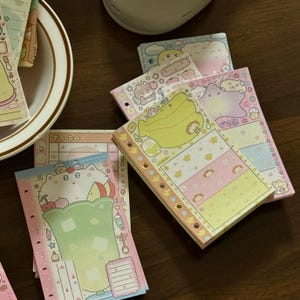 Puede incluir: Una colección de blocs de notas ilustrados y coloridos con varios diseños, incluyendo un plátano, un gato con un helado y otros personajes lindos. Los blocs son de colores pastel y tienen bordes decorativos y texto.