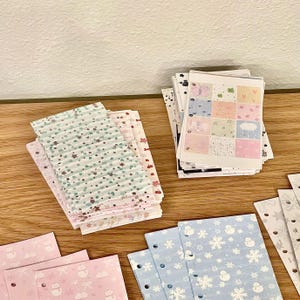 Könnte beinhalten: Eine Sammlung dekorativer Papierbögen mit verschiedenen Mustern. Die Bögen zeigen Designs wie Sterne, Schneeflocken, Schmetterlinge und andere niedliche Illustrationen. Die Farbpalette umfasst Pastellrosa, Blau, Grün und Weiß, was eine skurrile Ästhetik schafft.