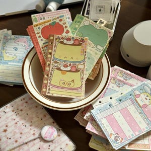 Puede incluir: Una colección de artículos de papelería coloridos, que incluyen cuadernos y blocs de notas, con lindos personajes de dibujos animados e ilustraciones de comida. Los artículos están dispuestos en un cuenco blanco y sobre una superficie de madera.