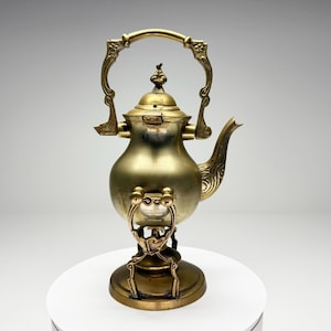 May include: A vintage brass teapot with ornate detailing. The teapot features a curved spout, a decorative handle, and a small base. The design includes floral and scrollwork patterns. The teapot is displayed on a white surface.