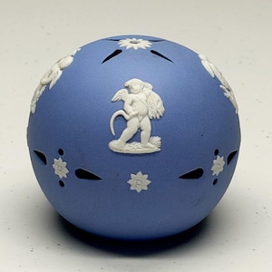 Può includere: Un pomander sferico in ceramica blu con figure in rilievo bianco di cherubini alati e motivi floreali. La superficie è punteggiata da piccoli intagli regolarmente distanziati e perforazioni decorative a forma di stella.