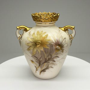 Puede incluir: Jarrón de cerámica color crema con detalles dorados y diseño floral. El jarrón tiene dos asas y un borde dorado decorativo. Las flores están pintadas en tonos amarillos, morados y marrones.