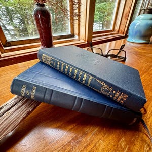May include: Two navy blue hardcover books stacked on a wooden surface. The top book is titled The Divine Comedy of Dante Alighieri with gold lettering on the spine. The bottom book is titled Poems of Tennyson with gold lettering on the spine.