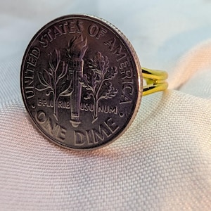 May include: A handcrafted ring featuring a United States one dime coin. The coin is set in a gold-colored band. The coin's details, including the words "ONE DIME" and "UNITED STATES OF AMERICA", are visible.