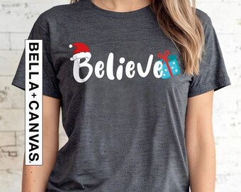 May include: A dark gray t-shirt with the word "Believe" in white lettering. A red Santa hat and a blue and red gift box are next to the word.