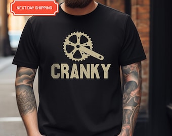 May include: Black t-shirt featuring a bicycle crankset graphic above the word "CRANKY" in a distressed font. The design is in a light tan color. The shirt is short-sleeved.