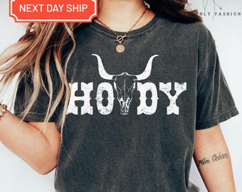 May include: Dark gray t-shirt with the word "HOWDY" in distressed white lettering, featuring a bull skull graphic. The shirt has a crew neck and short sleeves. The text "NEXT DAY SHIP" is in a red box in the upper left corner.