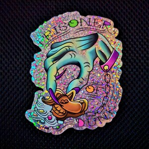 May include: A holographic sticker with a glittery background featuring a blue hand with a pink cuff and chain, holding a brown object. The text "Prisoner of Duty" is written in a rainbow font around the hand.