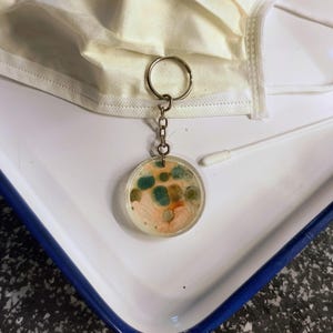 Puede incluir: Un llavero redondo con un colgante de resina transparente. El colgante presenta un patrón colorido de verde, naranja y blanco, que se asemeja a una placa de Petri. El llavero está unido a un anillo y una cadena plateados. Una mascarilla blanca y un hisopo están en el fondo.