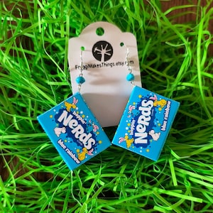 May include: A pair of handmade earrings featuring a blue Nerds candy box design. The earrings have silver hooks and turquoise beads. The text "Nerds" and "blue rainbow" are visible on the earrings. The background is green.