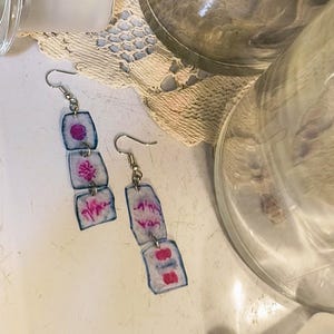 May include: Handmade earrings with a unique design. Each earring features three rectangular, translucent pieces connected by small silver links. The pieces have a white background with blue edges and pink abstract designs. The earrings hang from silver hooks.
