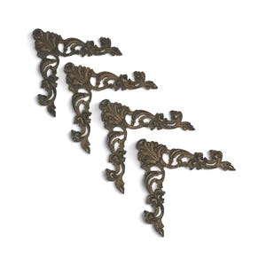 May include: Four antique bronze-colored corner brackets with ornate, scrolling floral designs. The decorative metal pieces are arranged diagonally against a white background, showcasing their intricate details and vintage aesthetic.