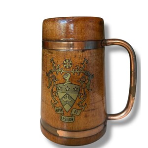 May include: Wooden beer stein with a copper handle and accents. The stein features a decorative crest with the words "Alpha Pi Epsilon". The wood has a warm, natural tone, and the copper has a vintage look.