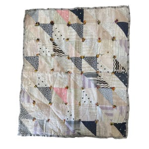 May include: Handmade patchwork quilt featuring a variety of fabrics in cream, pink, navy, and gray. The quilt is composed of triangular patches with dotted, striped, and patterned designs. Small, colorful embellishments are sewn at the intersections of the triangles.