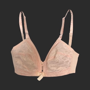 May include: A light pink bra with adjustable straps. The bra features a delicate lace overlay on the cups and a small bow detail at the center. The design includes a front closure and a soft, feminine aesthetic.