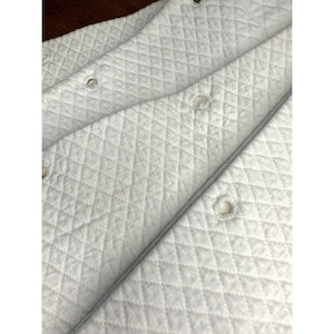 May include: Close-up of a white quilted fabric with a diamond pattern. The fabric has several buttonholes and appears to be part of a larger item. The texture is soft and the color is a clean white.