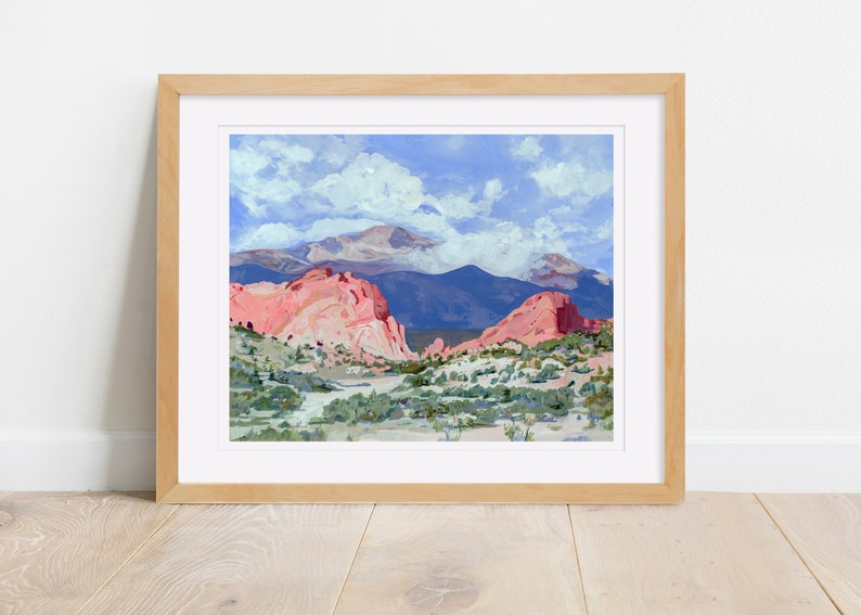 Op de afbeelding: Een ingelijst schilderij van een landschap met rode rotsformaties op de voorgrond en een bergketen op de achtergrond. De lucht is blauw met witte wolken.