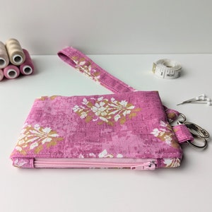 May include: A pink zippered pouch with a floral pattern and a gold strap. The pouch has a zipper closure and a key ring.