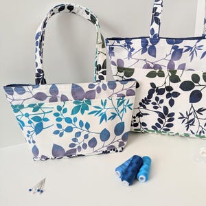 May include: Two tote bags with a white background and a blue and green leafy pattern. The smaller bag is in the foreground and the larger bag is in the background. There are spools of blue thread and sewing pins on the white surface.