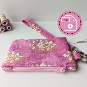 Puede incluir: Un estuche con cremallera con estampado floral rosa con una correa de muñeca a juego. El estuche tiene un diseño floral blanco y dorado. Un icono de descarga PDF está en la esquina superior derecha. También se ven suministros de costura.