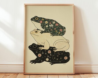 May include: A framed art print featuring three stylized frogs stacked on top of each other. The top and bottom frogs are black with floral patterns, while the middle frog is beige. The background is a light beige color.