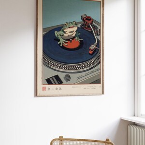 May include: A framed art print featuring a green frog on a blue record on a turntable. The frog is centered, with a red disc beneath it. The turntable is silver, and the background is a light blue. The print has Japanese text.