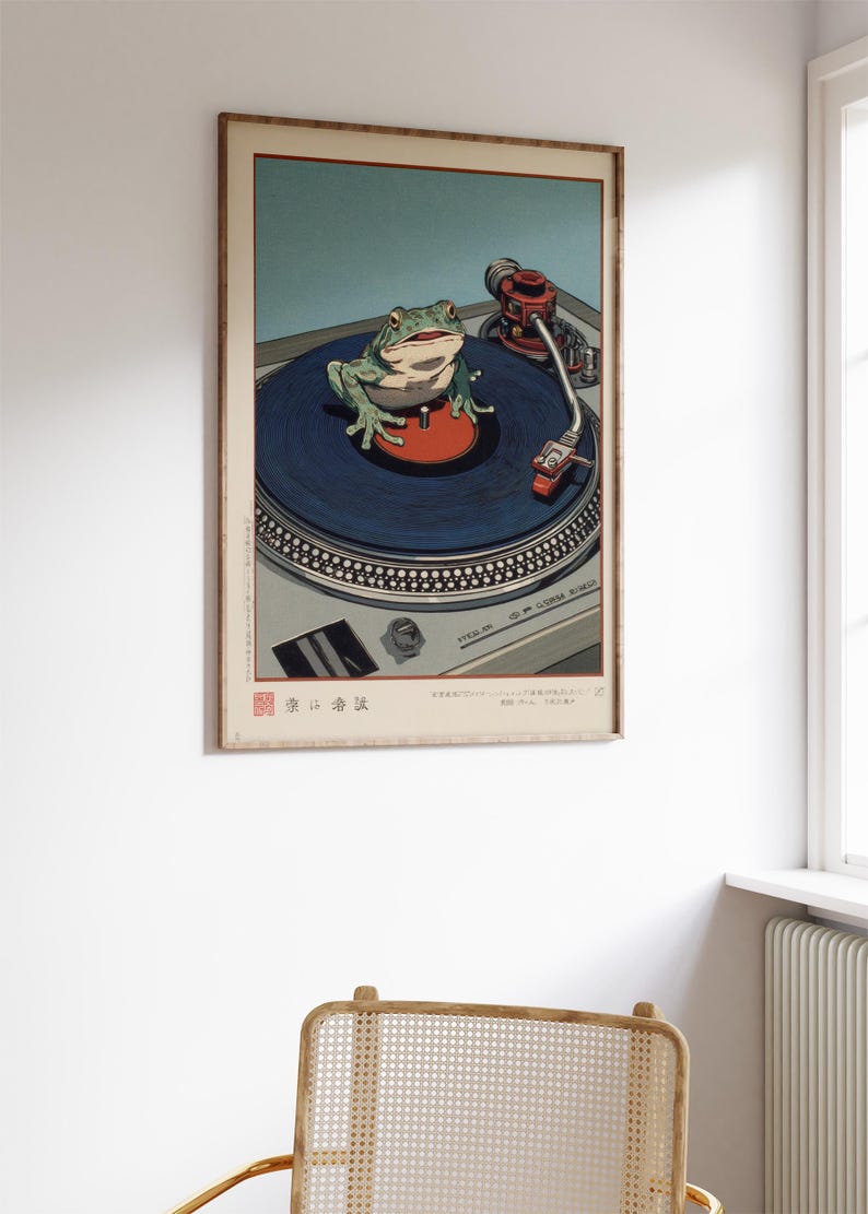 May include: A framed art print featuring a green frog on a blue record on a turntable. The frog is centered, with a red disc beneath it. The turntable is silver, and the background is a light blue. The print has Japanese text.