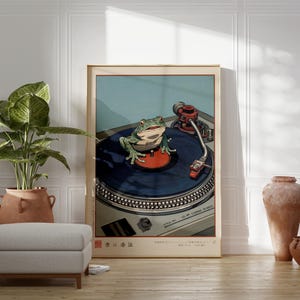 May include: A framed art print featuring a green frog on a record player. The frog is centered on the blue vinyl record, with the turntable's details visible. The background is a light blue, with Japanese text at the bottom.