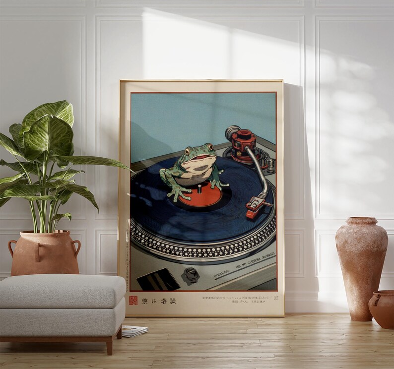 May include: A framed art print featuring a green frog on a record player. The frog is centered on the blue vinyl record, with the turntable's details visible. The background is a light blue, with Japanese text at the bottom.