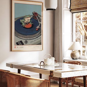 May include: A framed print depicts a green frog on a blue record player turntable. The artwork is in a room with a white table, rattan chairs, and a hanging paper lantern. The room has a window with a bamboo shade and a table lamp.