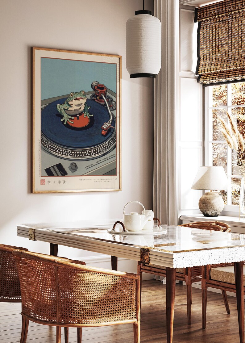 May include: A framed print depicts a green frog on a blue record player turntable. The artwork is in a room with a white table, rattan chairs, and a hanging paper lantern. The room has a window with a bamboo shade and a table lamp.