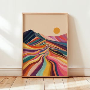 May include: A framed art print showing a colourful mountain landscape. The artwork depicts a winding path through the mountains, with vibrant hues of blue, green, yellow, orange, and pink. A yellow sun is in the upper right corner. The frame is made of light-coloured wood.
