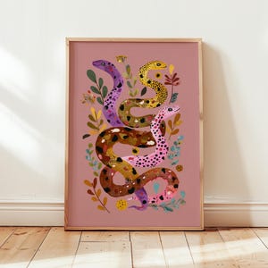 Könnte beinhalten: Gerahmter Kunstdruck mit drei bunten Schlangen, die sich mit floralen Elementen auf einem staubrosafarbenen Hintergrund verweben. Die Schlangen sind in Lila, Gelb, Braun und Rosa mit detaillierten Mustern dargestellt. Der Rahmen ist aus Naturholz.