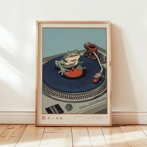 May include: Framed art print featuring a green frog on a blue record player. The frog is centered on the record, with the needle arm and other components of the turntable visible. The background is a light blue, and the frame is wood.