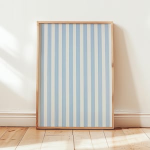 May include: A framed artwork featuring vertical stripes in light blue and off-white. The artwork is encased in a natural wood frame and leans against a white wall on a wooden floor. The overall aesthetic is clean and minimalist.