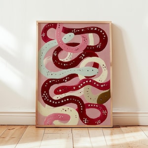 Puede incluir: Una impresión artística enmarcada con un diseño abstracto de serpientes entrelazadas. Las serpientes están pintadas en tonos de rojo, rosa y crema, con acentos de azul claro y marrón. El fondo es rosa suave y la obra de arte está en un marco de madera.