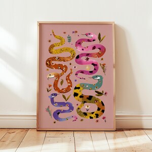 Könnte beinhalten: Ein gerahmter Kunstdruck mit Illustrationen von Schlangen in leuchtenden Farben und Mustern, wie Gelb, Orange, Rosa und Blau, auf rosa Hintergrund. Das Kunstwerk ist mit floralen Akzenten verziert.
