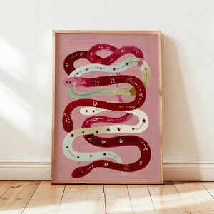 Pode incluir: Uma impressão artística emoldurada com uma ilustração estilizada de cobras entrelaçadas. As cobras são pintadas em tons de vermelho, branco e verde claro, com pequenos detalhes florais sobre um fundo rosa. A obra de arte é apresentada numa moldura de madeira.