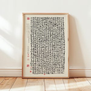 May include: A framed artwork featuring calligraphy in black ink on a cream-colored background. The calligraphy is dense and detailed, with numerous characters arranged vertically. Red stamps are visible in the corners and along the left side. The frame is made of light-colored wood.
