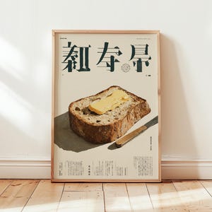 Puede incluir: Impresión enmarcada con texto japonés, que representa una rebanada de pan con mantequilla y un cuchillo. El pan es marrón con una porción de mantequilla amarilla. El cuchillo es marrón con una hoja plateada. El marco es de madera.