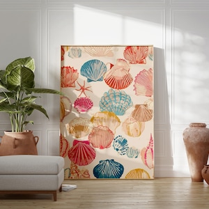 May include: A framed artwork featuring a repeating pattern of colorful seashells in shades of red, blue, pink, and yellow on a cream background. The artwork is leaning against a white wall, with a plant and ottoman in the foreground.