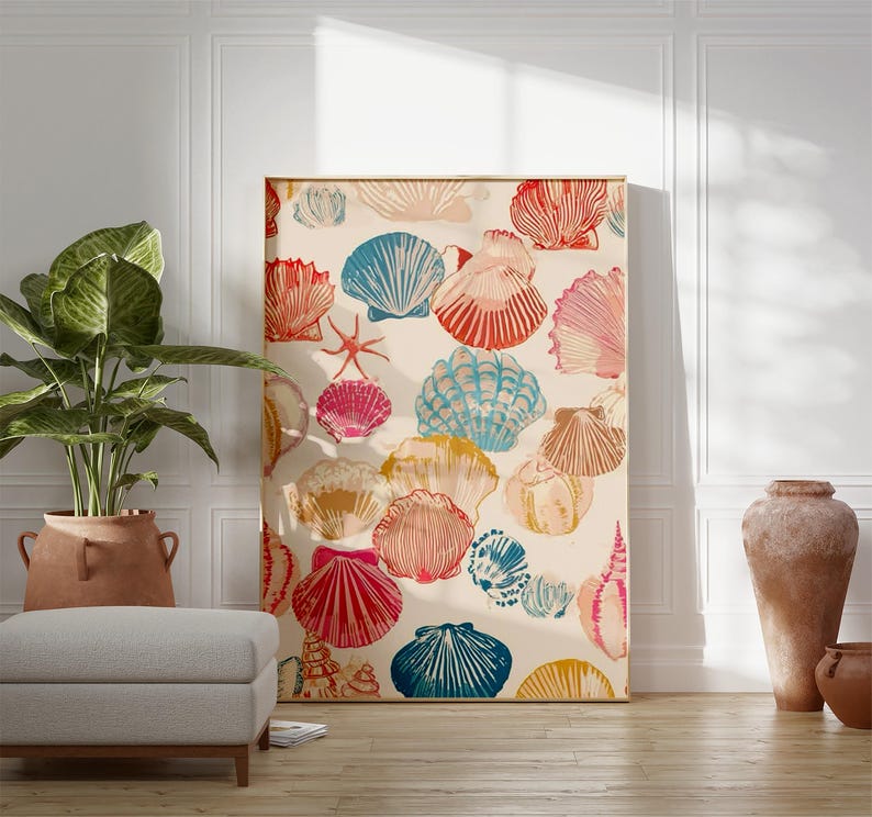 May include: A framed artwork featuring a repeating pattern of colorful seashells in shades of red, blue, pink, and yellow on a cream background. The artwork is leaning against a white wall, with a plant and ottoman in the foreground.