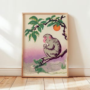 May include: Framed vintage print of a Japanese macaque monkey perched on a tree branch. The artwork features a gray monkey with a pink face, green leaves, and an orange fruit against a purple and cream background. The print is in a wooden frame.