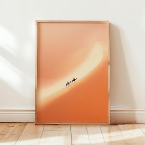 Könnte beinhalten: Gerahmter Druck einer Wüstenlandschaft mit zwei Kamelen und Reitern, die eine Sanddüne hinaufsteigen. Das Bild hat eine warme Farbpalette aus Orange und Beige, in einem Holzrahmen.