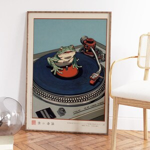 May include: Framed art print featuring a green frog on a blue record, positioned on a silver turntable. The artwork has a vintage aesthetic with a red tone and Japanese text at the bottom. The background is light blue.